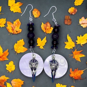 Może przedstawiać: Para kolczyków wiszących z zawieszką w kształcie srebrnej czaszki i czarnymi i fioletowymi koralikami. Kolczyki wiszą na srebrnych haczykach i są umieszczone na tle jesiennych liści.