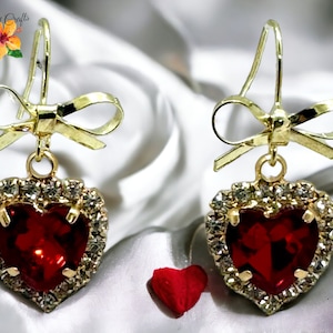 Może przedstawiać: Złote kolczyki z czerwonym sercem ozdobionym przezroczystymi cyrkoniami i złotą kokardką.