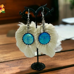 May include: A pair of white tassel earrings with a blue face cameo pendant. The earrings are hanging from a black earring stand.