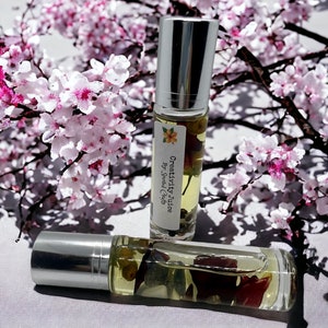 May include: Two clear glass roller bottles with silver caps filled with a yellow liquid and dried flowers. The bottles are labeled "Creativity Juice by Jewel Crag" and are sitting on a white surface.