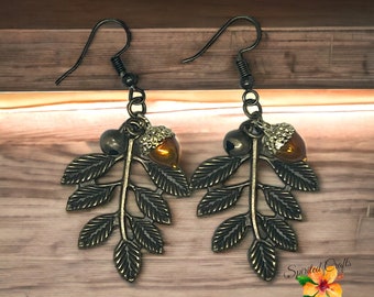 Puede incluir: Un par de pendientes colgantes de bronce con forma de bellota y hoja. Los pendientes presentan una rama delicada con varias hojas y dos bellotas con detalles dorados.