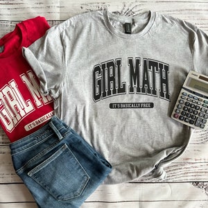 Puede incluir: Una camiseta gris con el texto "Girl Math" en letras negras en bloque y el texto "It's basically free" en letras negras más pequeñas. La camiseta está doblada y colocada sobre una superficie de madera blanca con un par de jeans azules y una calculadora.