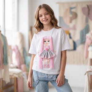 May include: White t-shirt with a doll design. The doll has long pink hair, a gray and pink dress, and pink shoes. The shirt is worn with blue denim overalls. The doll has a flower on its dress.