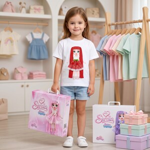 Peut inclure: Une jeune fille porte un t-shirt blanc avec un motif de poupée et tient une boîte rose avec une illustration de poupée. La boîte porte l'inscription "Sirella Dream Princess". D'autres boîtes et vêtements sont visibles en arrière-plan.