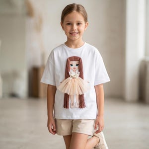 May include: A white t-shirt with a 3D doll design. The doll has long brown hair, a peach-coloured tutu, and a floral bodice. The doll's face has blue eyes. The shirt is worn by a young person.