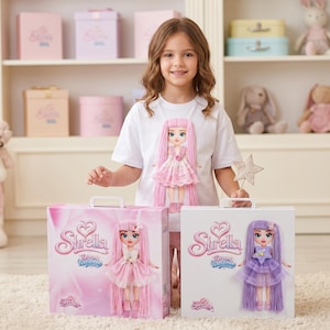 Könnte beinhalten: Ein weißes T-Shirt mit einer Grafik einer Puppe mit langem rosa Haar und rosa Kleid, präsentiert neben zwei rechteckigen Shrella Dream Wardrobe Spielzeugkoffern, einer in Rosa und einer in Weiß, jeweils mit Puppenabbildung.
