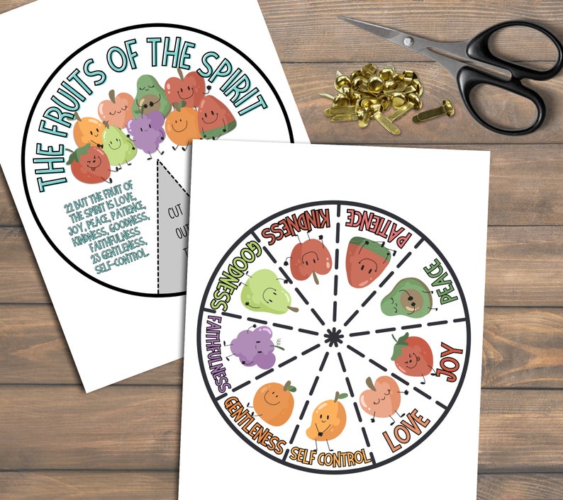 May include: Two circular educational printables with the text "The Fruits of the Spirit" and a list of virtues. One features illustrated fruits with faces, and the other is a wheel with virtues. Scissors and fasteners are also visible.