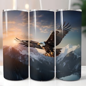 May include: A stainless steel tumbler with a design of a bald eagle flying over a mountain range with a sunset in the background.