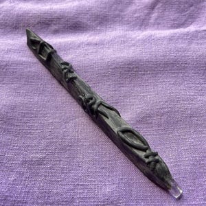 Może przedstawiać: Ciemnoszara różdżka ze skomplikowanymi, wypukłymi czarnymi detalami. Różdżka ma spiczastą końcówkę i jest ozdobiona wirującymi wzorami przypominającymi winorośl. Tło to fioletowa tkanina.