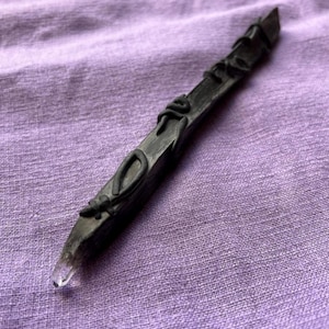 Może przedstawiać: Ciemnoszara, ręcznie wykonana różdżka z przezroczystą kryształową końcówką. Różdżka jest ozdobiona wypukłymi, czarnymi elementami dekoracyjnymi na całej długości. Tło to fioletowa tkanina.