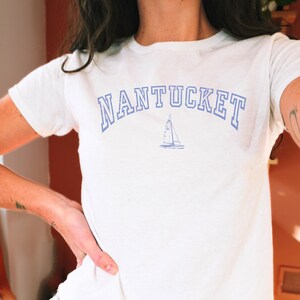 Könnte beinhalten: Ein weißes kurzärmeliges T-Shirt mit dem Wort NANTUCKET, das in einer blauen, bogenförmigen Schrift im College-Stil gedruckt ist. Unter dem Text befindet sich eine kleine blaue grafische Abbildung eines Segelboots. Das T-Shirt hat einen Rundhalsausschnitt.