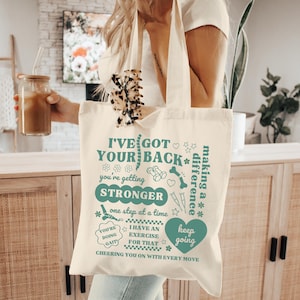 May include: Cream-colored tote bag with teal text that reads "I've Got Your Back" and other supportive phrases. The bag features illustrations of bones, hearts, and crutches. A woman is holding the bag, along with a glass of iced coffee.
