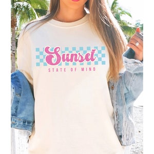 May include: A white t-shirt with a pink and blue checkered design and the text "Sunset State of Mind".