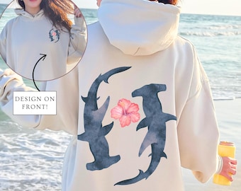 May include: A cream-colored hoodie with a design on the back featuring two hammerhead sharks and a pink hibiscus flower. The hoodie has a large hood and a relaxed fit.