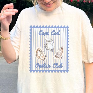 Puede incluir: Camiseta color crema con un diseño de sello azul. El diseño presenta las palabras "Cape Cod Oyster Club" e ilustraciones de tres ostras. El fondo tiene rayas azules.