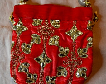 Puede incluir: Una bolsa de cordón roja con un asa de cuentas doradas y bordados dorados. La bolsa está decorada con un patrón floral.