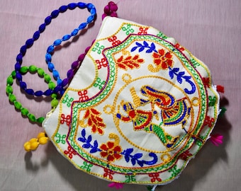 Puede incluir: Una bolsa de tela blanca con bordados coloridos que representan una escena de dos figuras bailando. La bolsa tiene una correa de cuentas con cuentas azules, verdes y moradas.