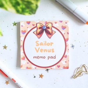 May include: A pink and orange memo pad with a Sailor Venus design. The pad features a large circle with the text "Sailor Venus memo pad" and a drawing of a bow with a heart in the centre.