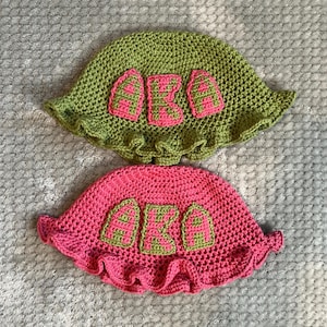 May include: Two handmade crochet hats. One is green with pink letters spelling "AKA", and the other is pink with green letters spelling "AKA". Both hats have a ruffled brim.