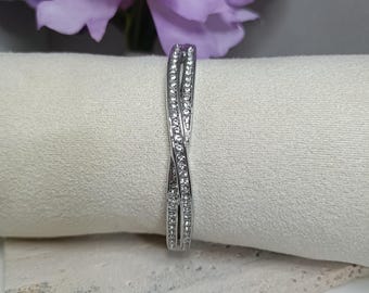 May include: A silver-colored bracelet with a twisted design, encrusted with small, clear gemstones. The bracelet is displayed on a white surface, with a blurred purple flower in the background.
