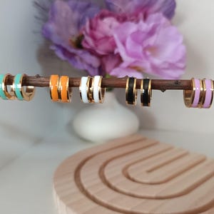 May include: A selection of gold hoop earrings with coloured enamel stripes. The earrings are displayed on a wooden branch, showcasing colours including teal, orange, white, black, and lavender. The background features flowers and a wooden display.