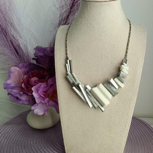 May include: A silver-toned necklace with geometric shapes and white rectangular accents. The necklace is displayed on a beige bust, with a background of purple flowers and decorative feathers. The necklace has a modern, abstract design.