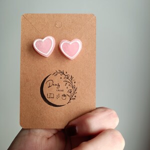 May include: A pair of pink heart-shaped earrings with white outlines. The earrings are displayed on a brown card with the text "Dainty Grove" in black.
