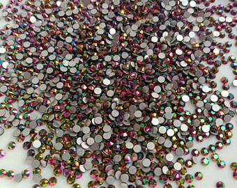 May include: A close-up view of a collection of iridescent rhinestones. The small, round gems display a range of colors, including pink, gold, and green, with a silver backing. The rhinestones are scattered on a white surface.