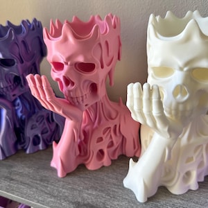 May include: Three skull-shaped planters in purple, pink, and white. Each planter features a skeletal figure with a crown-like top, a hand resting near the chin, and a textured, melting-effect surface design throughout the entire structure.