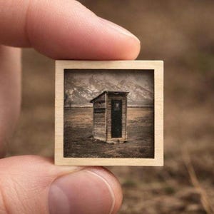 May include: A small square wooden frame containing a photographic print of a rustic wooden outhouse situated in a vast, dry field with a mountain range visible in the background. The image features muted brown, tan, and grey tones throughout.