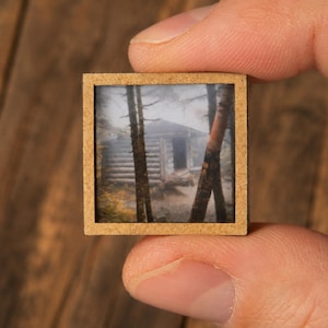 May include: A small square wooden frame containing a printed image of a log cabin nestled among tall trees in a misty forest. The frame has a light brown, textured finish and holds the landscape scene securely within its thin, flat border.
