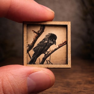 May include: A miniature artwork featuring a black bird perched on a branch, set within a small, square wooden frame. The bird is detailed with dark feathers, and the background is a sepia tone. The artwork is held between two fingers.