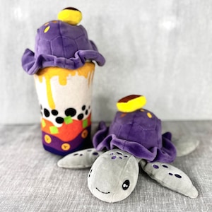 May include: Two plush toys, one shaped like a boba tea cup with a purple lid and the other shaped like a turtle with a purple shell and a grey head. The boba tea cup has a yellow and orange design with black boba pearls.