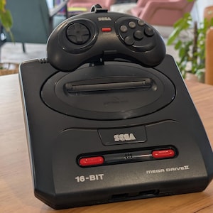 May include: A black Sega Mega Drive II video game console with a matching black controller resting on top. The console features the Sega logo, a cartridge slot, two red buttons, and the text 16-BIT and MEGA DRIVE II printed on the front surface.