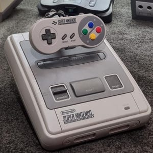 May include: A Super Nintendo Entertainment System console with a matching controller. The console is light grey with a darker grey centre section. The controller has a directional pad, select, start, and coloured buttons.