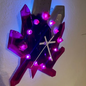 May include: A purple and white crystal-shaped wall clock with glowing pink lights. The clock has white hands and a white face.
