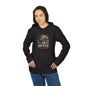 Könnte beinhalten: Ein schwarzer Kapuzenpullover mit einem Grafikdruck eines Adlers mit grünen, weißen und roten Federn, einem Fußball und dem Text MEXICO WORLD CUP 2026. Der Hoodie verfügt über lange Ärmel, eine Kängurutasche und eine Kapuze mit Kordelzug.