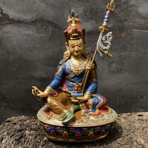 Könnte beinhalten: Eine handbemalte Statue von Padmasambhava, die im Schneidersitz auf einer Lotusbasis sitzt. Die Figur trägt ein blaues Gewand, goldenen Schmuck und eine Krone, hält einen Vajra in der rechten Hand und eine Schädelschale in der linken, mit einem Khatvanga-Stab.