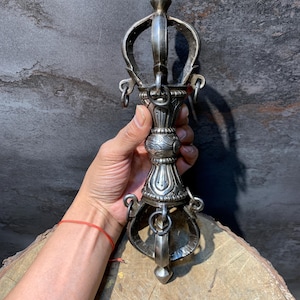 Op de afbeelding: Een zilverkleurige Vajra, een ritueel object met een centrale handgreep en sierlijke, gebogen tanden. Het object heeft ingewikkelde details en wordt tegen een donkere, getextureerde achtergrond gehouden. De Vajra is een symbool van het boeddhisme.