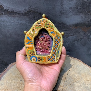 May include: A small, ornate religious icon held in a hand. The icon features a central carved figure in shades of red, framed by a decorative border with gold, orange, blue, and green patterns. The frame has gold accents and is held on a wooden surface.