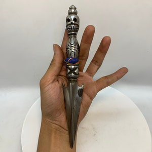 May include: A silver dagger with a skull-shaped handle and a blue and orange band around the handle. The dagger is held in a hand.