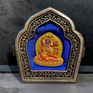 May include: A silver and blue shrine with a yellow and red painted deity inside. The shrine has a decorative floral design.