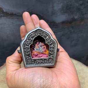 May include: A silver-colored, ornate reliquary with a detailed floral design. Inside, a small, colorful figure is visible. The reliquary is held in a hand, showcasing its size and intricate craftsmanship.