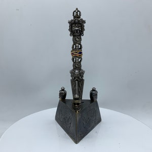 May include: A silver and black metal ritual dagger with a triangular base and intricate carvings. The dagger has a decorative handle with a crown and a colorful band.