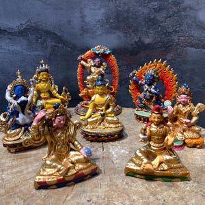 May include: A collection of seven small, colorful, gold-painted statues of deities. The statues are arranged on a wooden surface. Each statue is sitting on a base with intricate details. The statues are all different sizes and shapes, but they all have a similar style. The statues are all made of metal and are painted in bright colors.