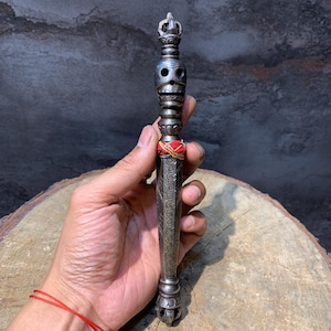 Pode incluir: Um objeto ritualístico prateado e ornamentado, com um design semelhante a um crânio e entalhes intrincados. O objeto tem um topo em forma de coroa e é envolto com fio vermelho e amarelo. É segurado contra um fundo escuro.