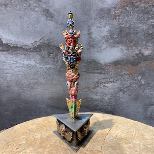 May include: A multi-tiered, brightly coloured sculpture featuring various figures and faces. It is mounted on a triangular base with decorative details. The sculpture incorporates red, blue, gold, and green hues, and is likely a decorative or religious artefact.