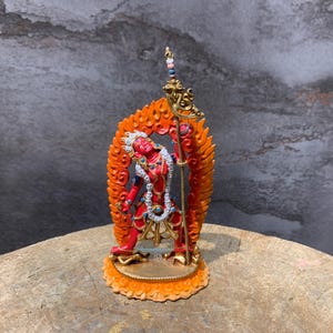 Op de afbeelding: Een gedetailleerd, kleurrijk beeld van een godheid met een rood lichaam, een witte hoofdtooi en sierlijke sieraden. De figuur houdt een staf vast en wordt omlijst door een levendige oranje en gele halo. De basis is een gedetailleerd, cirkelvormig ontwerp. Het beeld staat op een houten oppervlak.