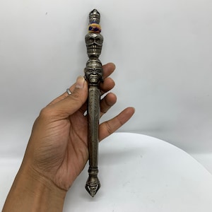 Könnte beinhalten: Ein silberfarbenes Metallgebetrad mit aufwendigen Schnitzereien und einem schädelartigen Design. Das Gebetsrad ist mit buntem Garn verziert.