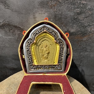 Könnte beinhalten: Ein goldfarbenes, rechteckiges Amulett mit einer zentralen Relief-Figur, eingefasst in einen silberfarbenen Rahmen mit detaillierten floralen Gravuren. Das Amulett ist auf gelbem Stoff befestigt und befindet sich in einem gelb-weinroten Klappetui.